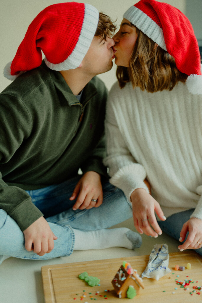 Expecting couple kiss as they finished decorating their gingerbread house with festive santa hats on their heads
